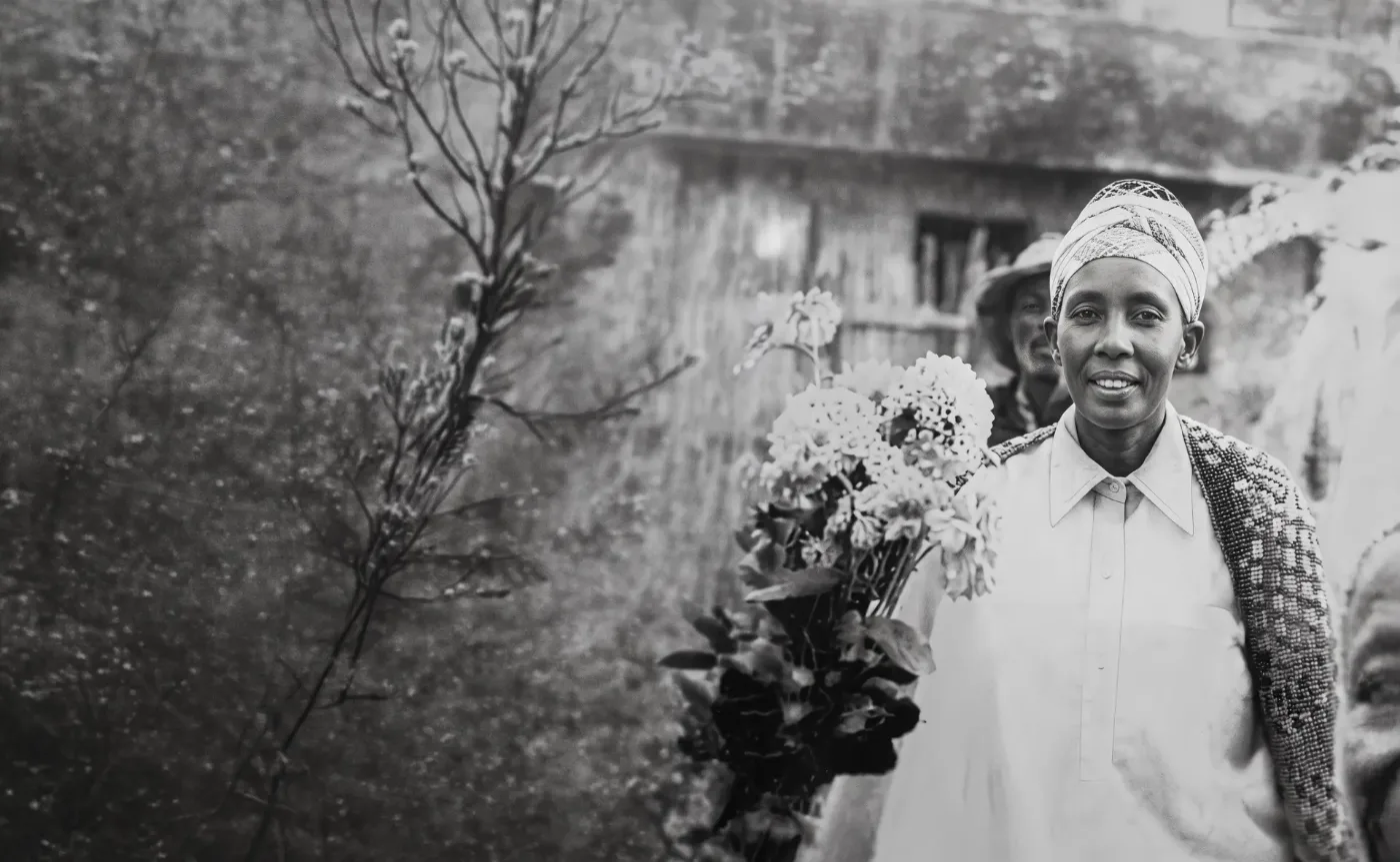 A smiling woman in a white blouse and headwrap holding a large bouquet of wildflowers before a timber building, archival black-and-white photograph
