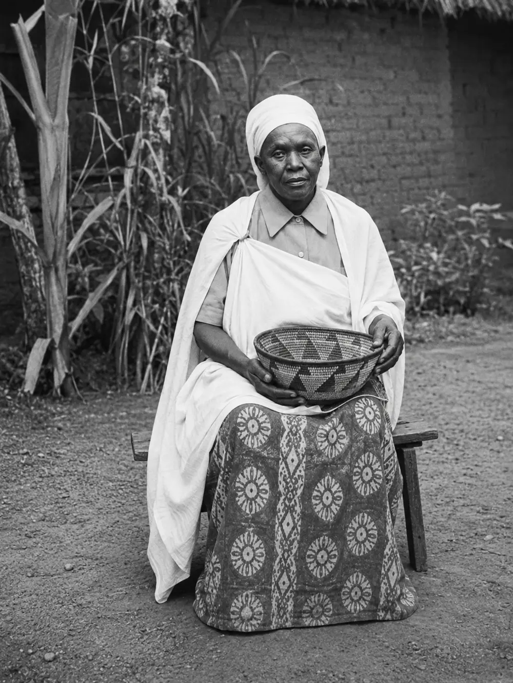 A woman seated on a wooden bench holding a woven basket, wearing a white wrap and a patterned skirt