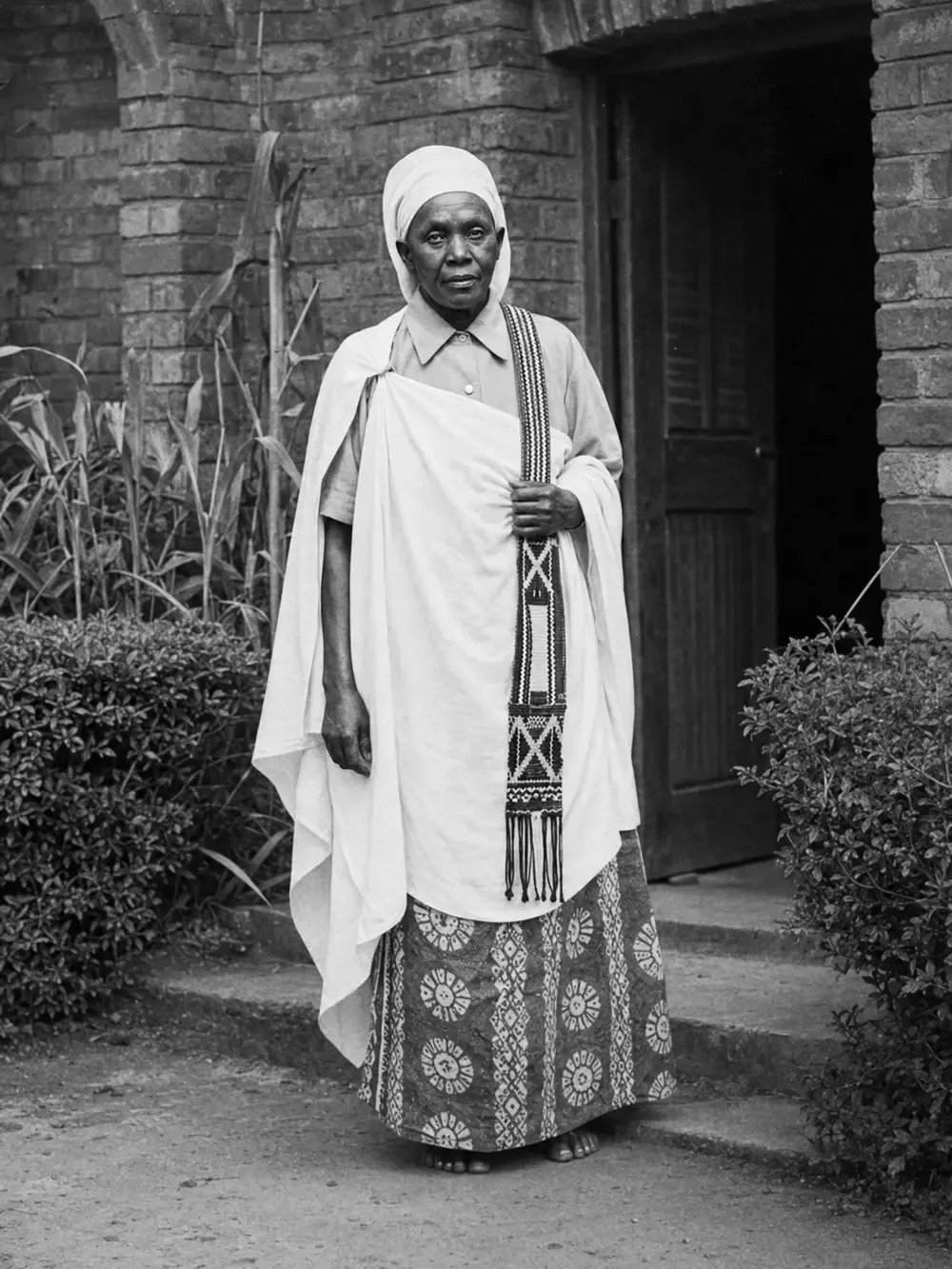 Portrait of a woman in a white wrap with a beaded sash, standing before a brick doorway